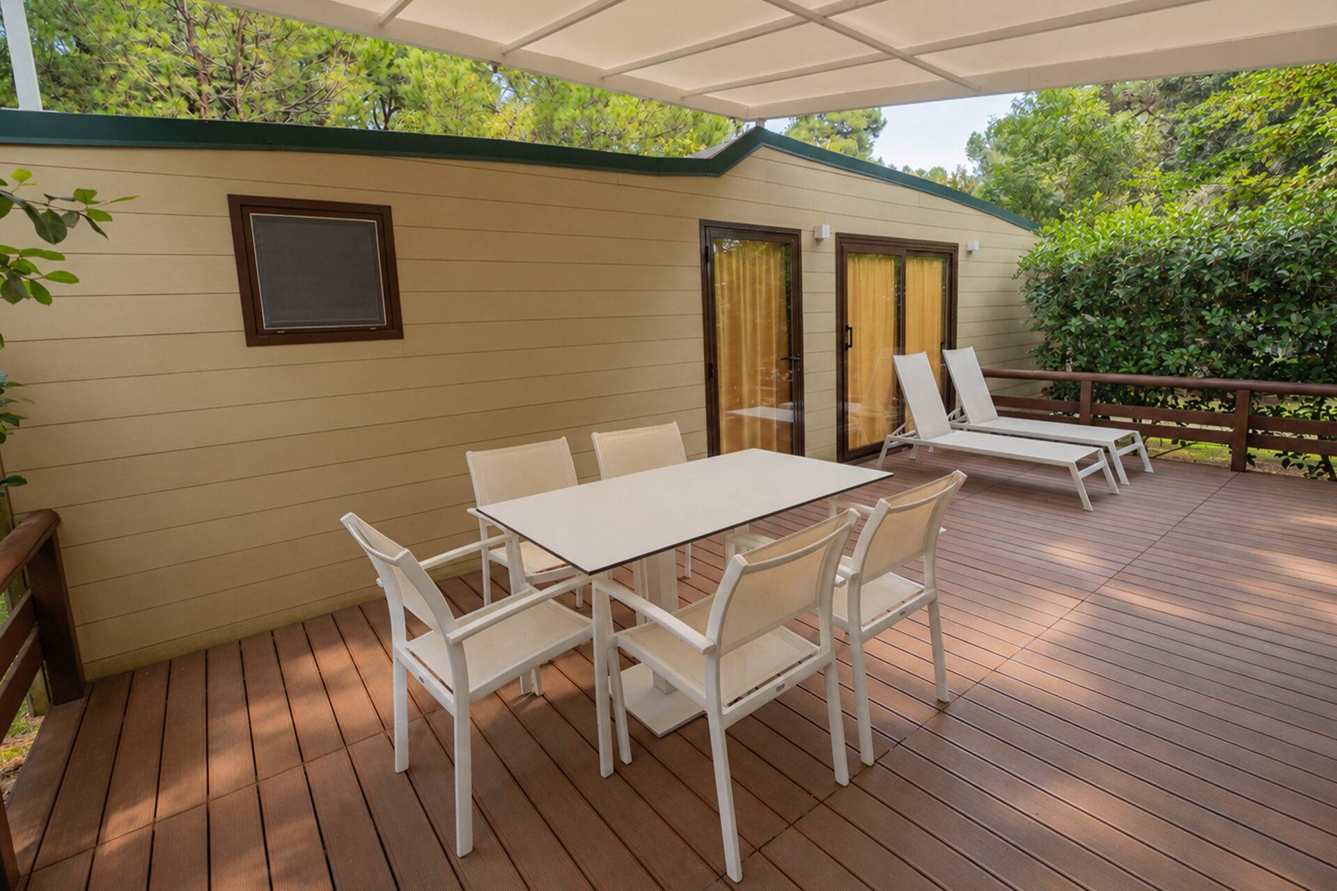 Terrazza coperta dello Chalet Natura Classic con tavolo, sedie e due lettini prendisole, circondata dal verde.