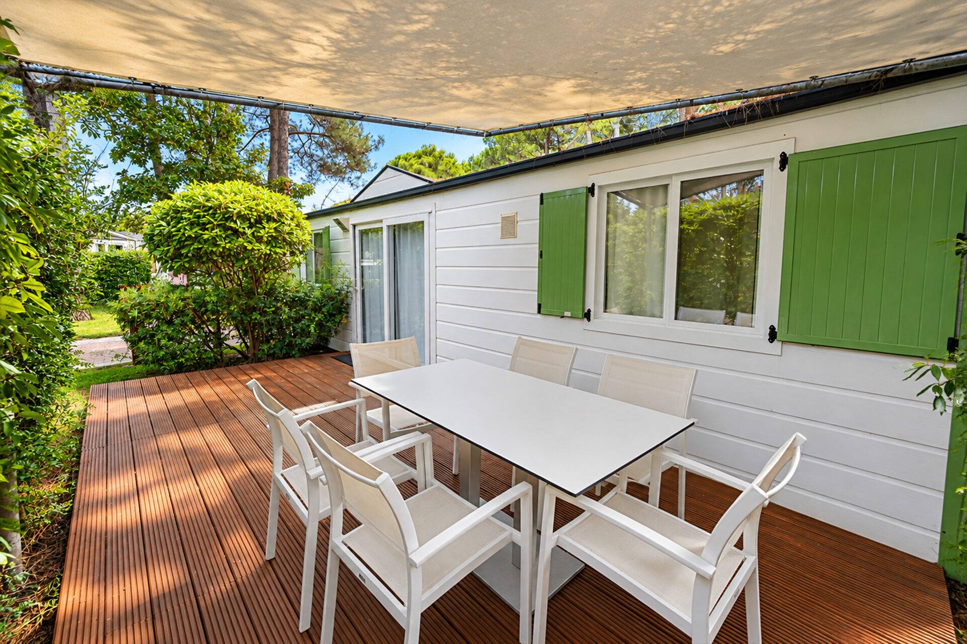 Terrazza in legno dello Chalet Sole Six Beds II con tavolo, sei sedie e telo ombreggiante, circondata da siepi e alberi.