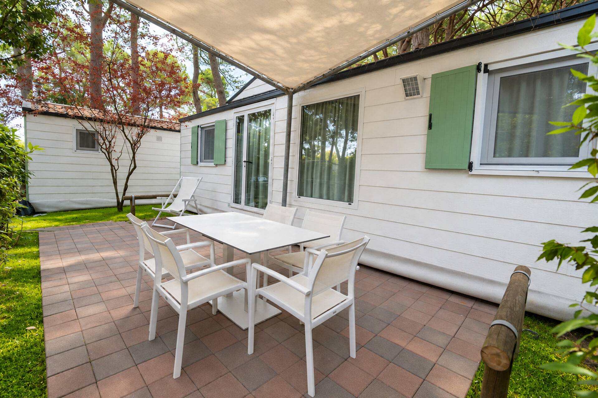 Veranda dello Chalet Sole Classic I con tavolo, sedie bianche e sdraio sotto il telo ombreggiante, circondata dal verde.