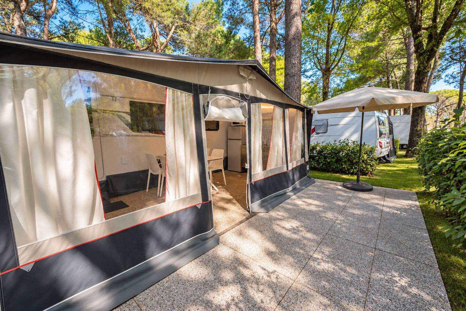 Veranda della Caravan Tabbert con pavimentazione esterna, ombrellone e siepi verdi nella pineta del Camping Village Dei Fiori.