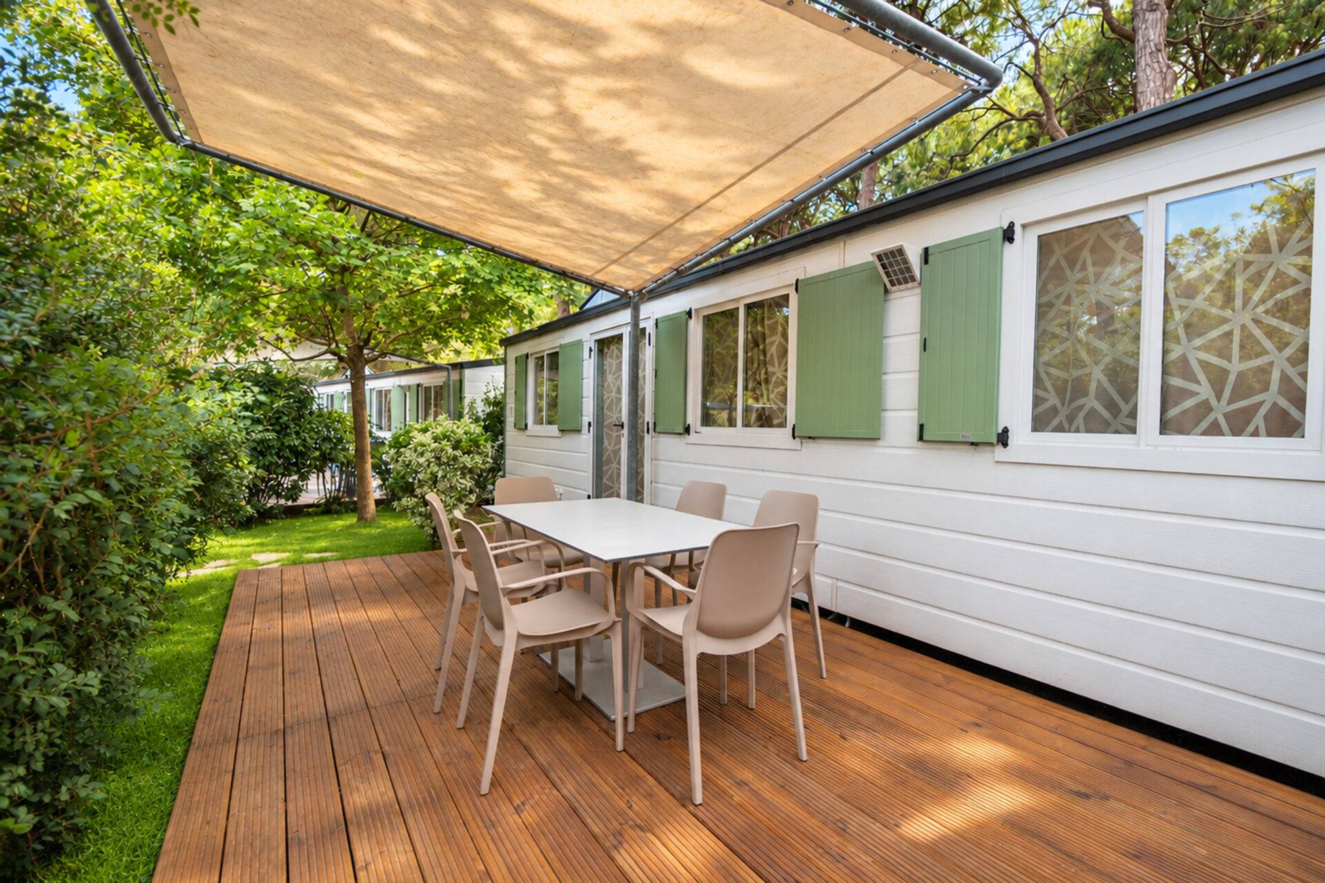 Terrazza in legno dello Chalet Sole Six Beds I con tavolo, sedie e telo ombreggiante, circondata dal verde.