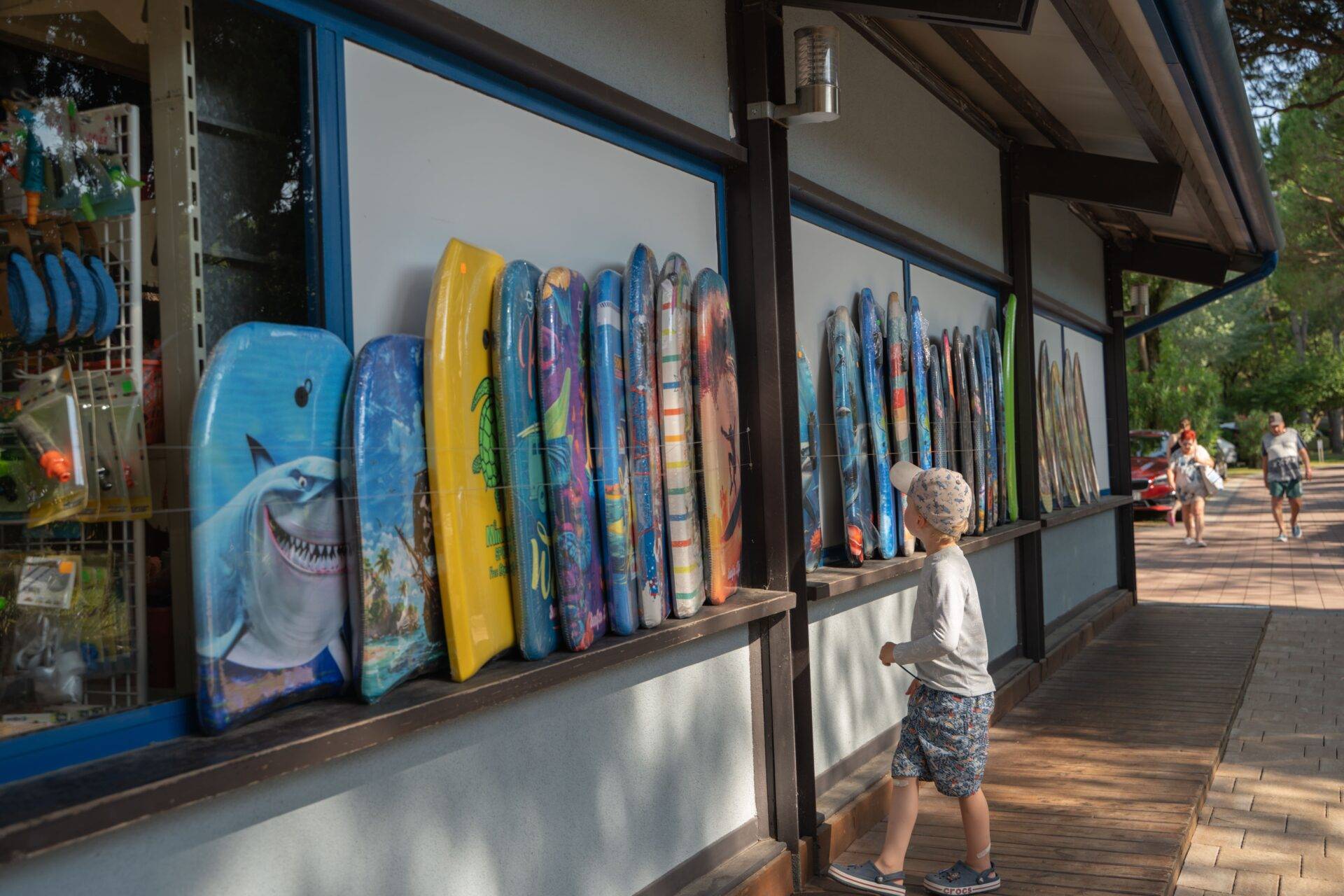 Bambino osserva bodyboard e tavole da mare esposte davanti al bazar del Camping Village Dei Fiori.