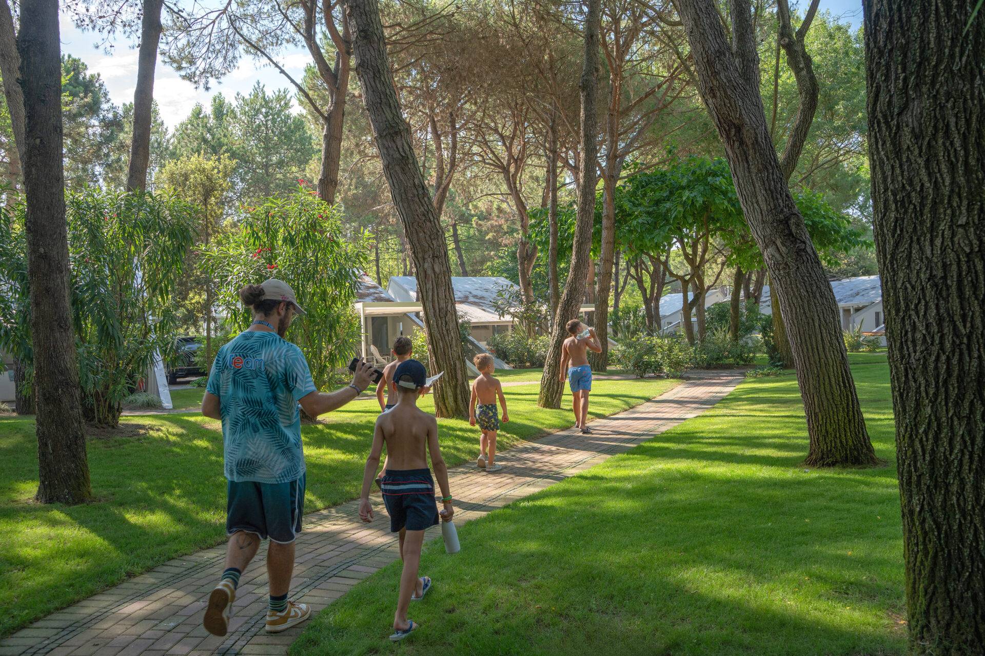 Animatore del Camping Dei Fiori accompagna un gruppo di ragazzi lungo un vialetto alberato durante un'attività del Maxi Club.