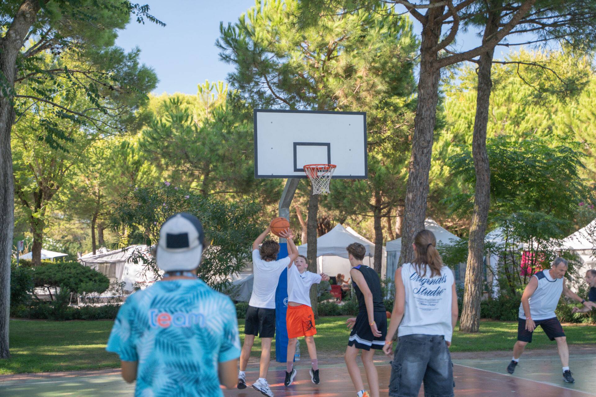 Ragazzi giocano a basket sul campo del Camping Dei Fiori, mentre un animatore osserva la partita tra i pini.