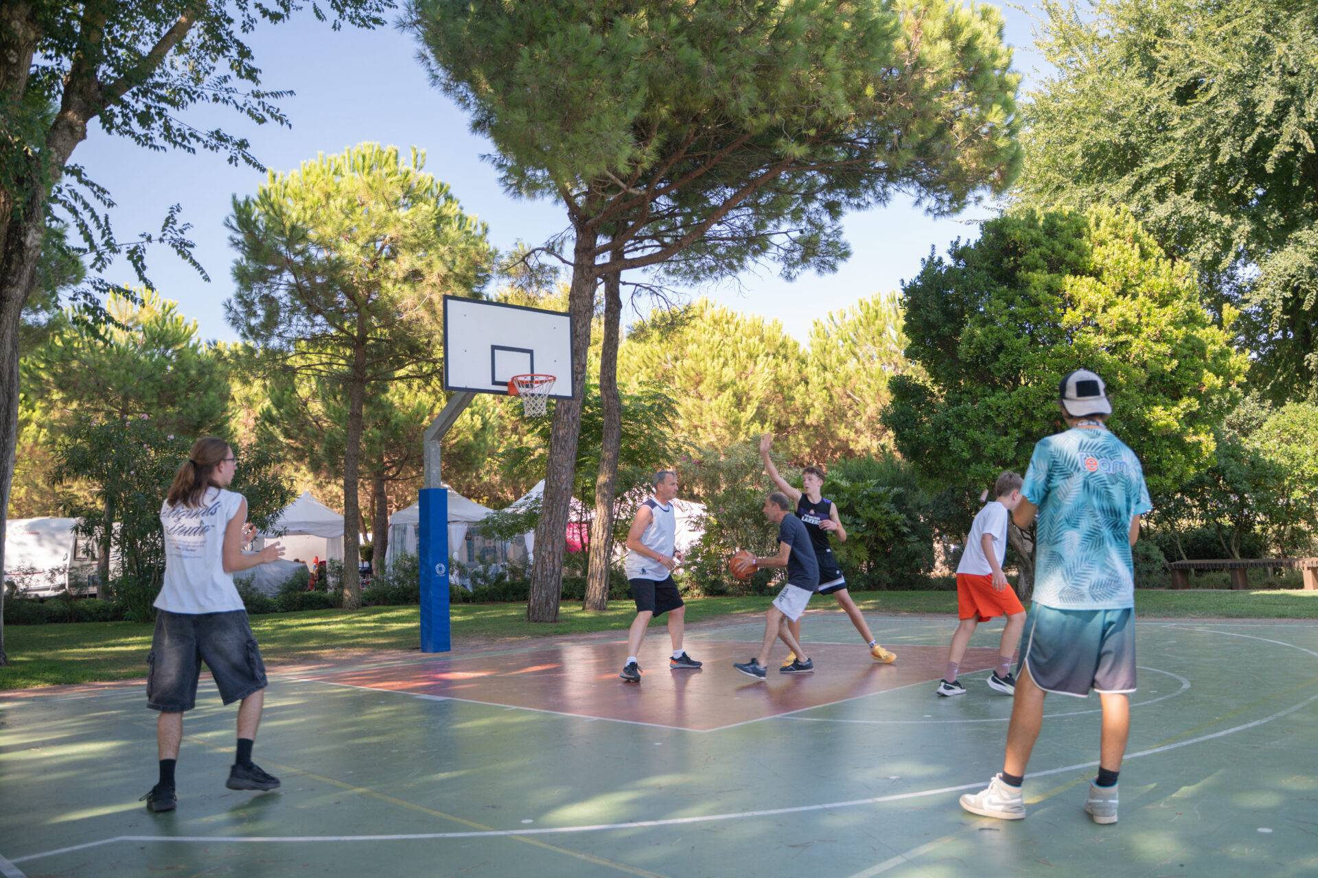 Gruppo di ragazzi e adulti gioca a basket sul campo all'aperto del Camping Village Dei Fiori, circondato da alberi.