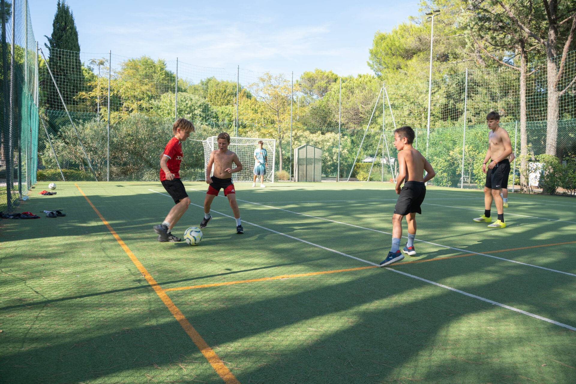 Ragazzi giocano a calcetto sul campo sportivo del Camping Village Dei Fiori, circondato da alberi e vegetazione.