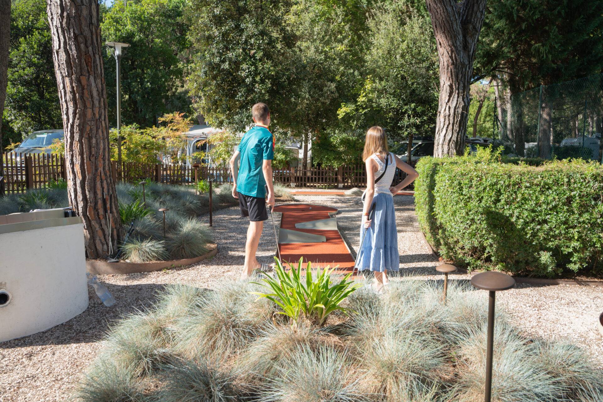 Due giovani giocano a minigolf su una pista rossa circondata da alberi e piante al Camping Village Dei Fiori.