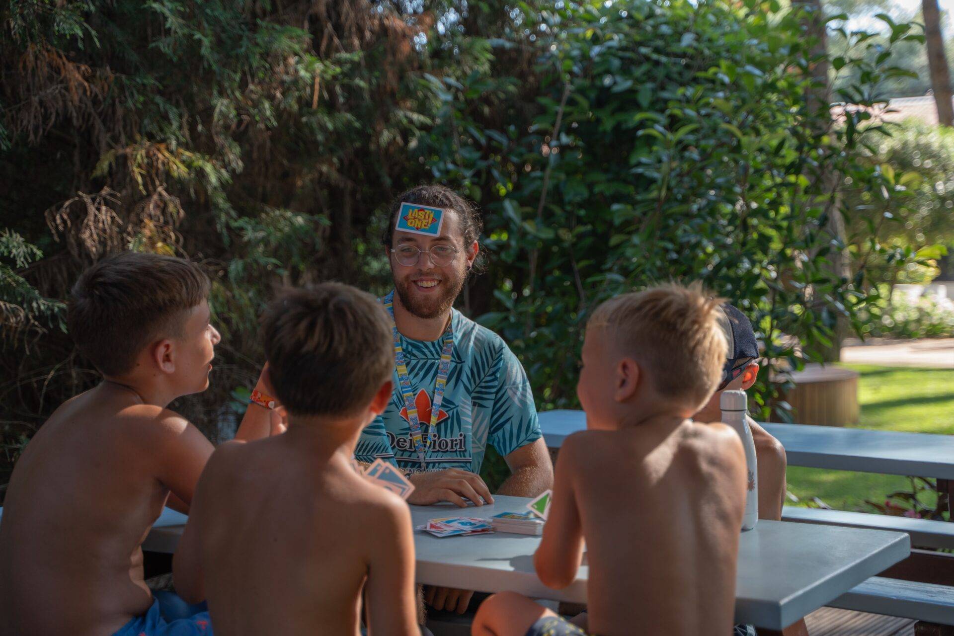 Animatore del Camping Dei Fiori gioca a carte con quattro ragazzi seduti a un tavolo all'aperto, circondati dal verde.
