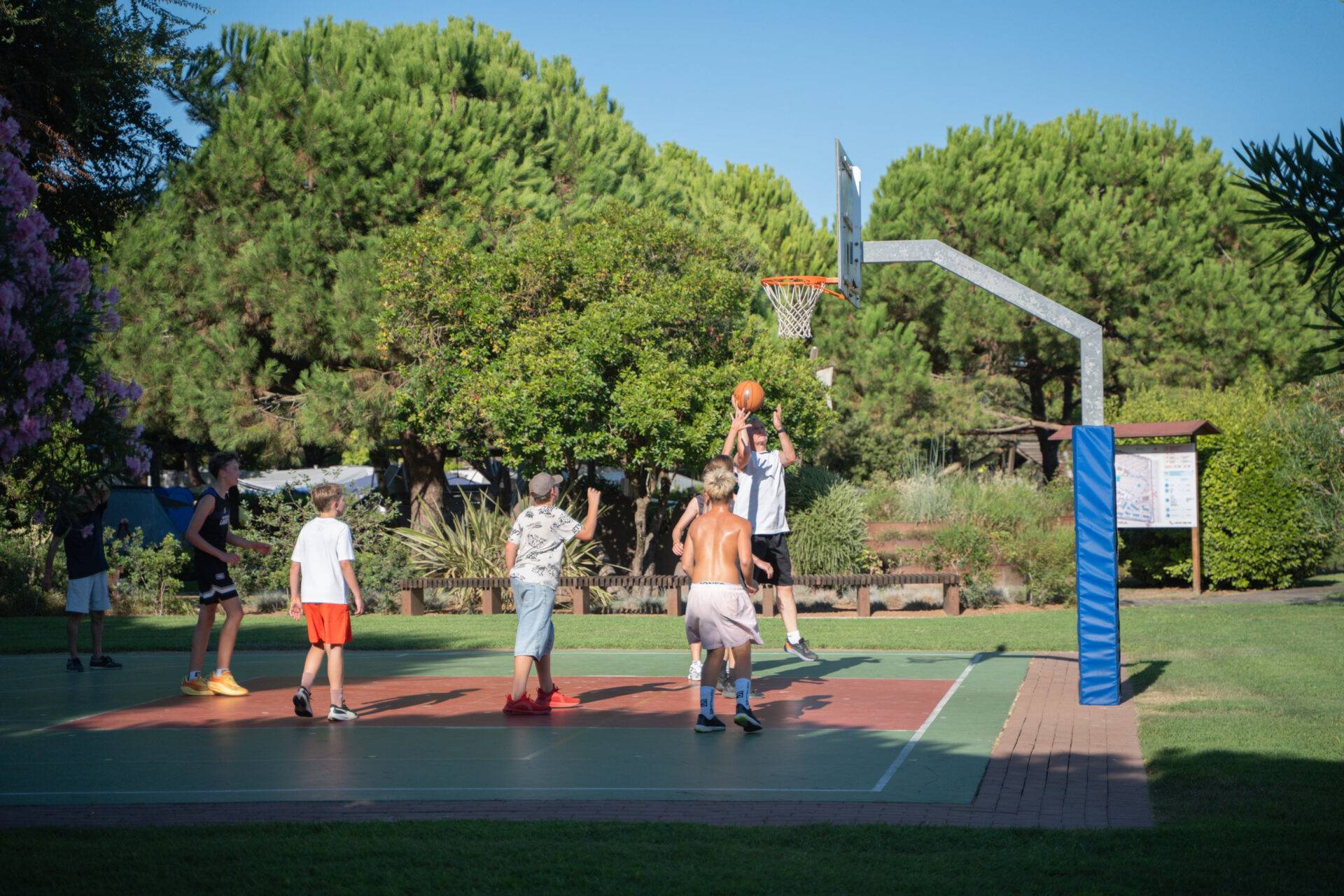 Gruppo di ragazzi gioca a basket sul campo all'aperto del Camping Dei Fiori, circondato da alberi e prati verdi.