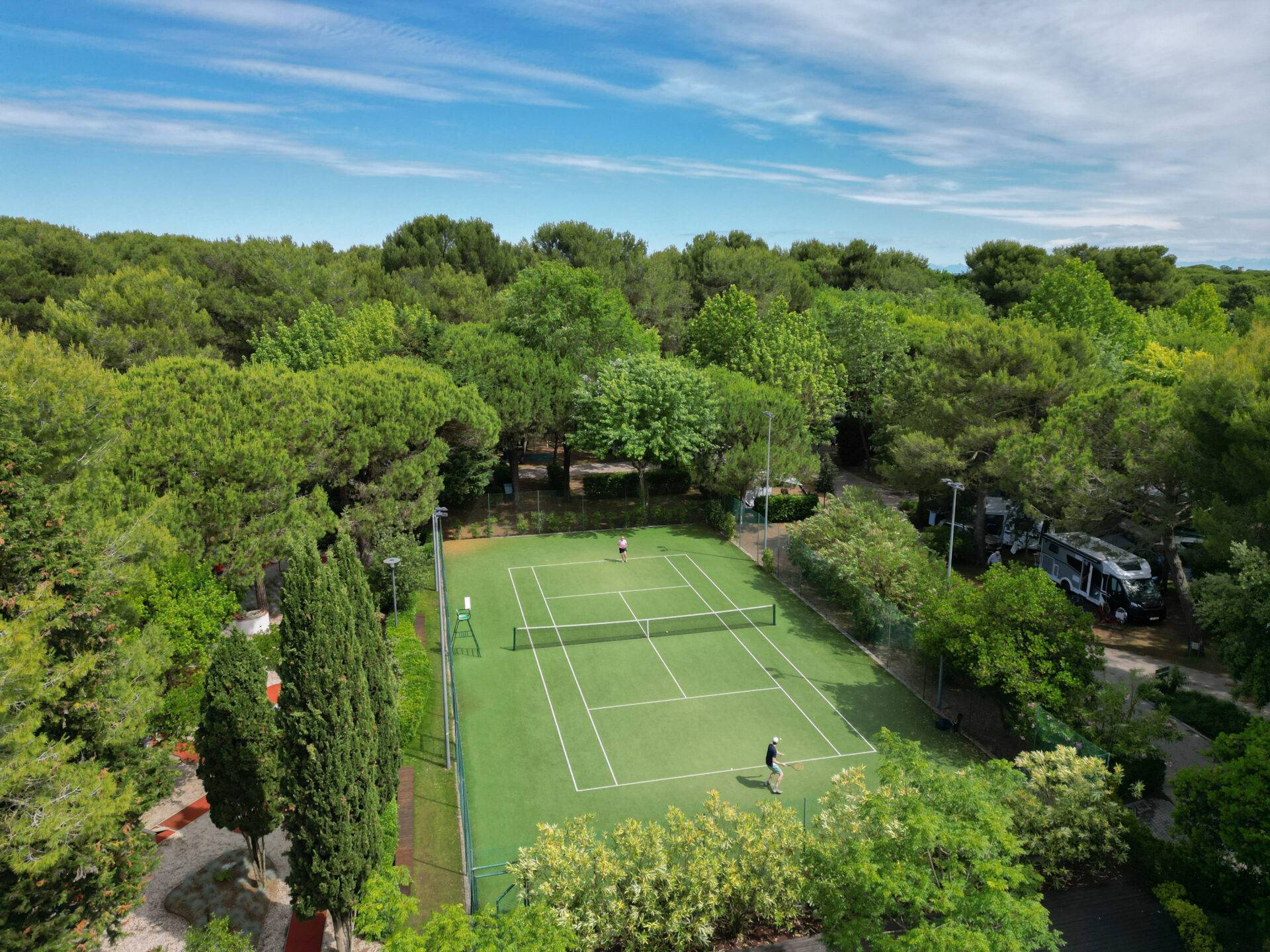 Due persone giocano a tennis sul campo immerso nel verde del Camping Village Dei Fiori a Cavallino-Treporti.