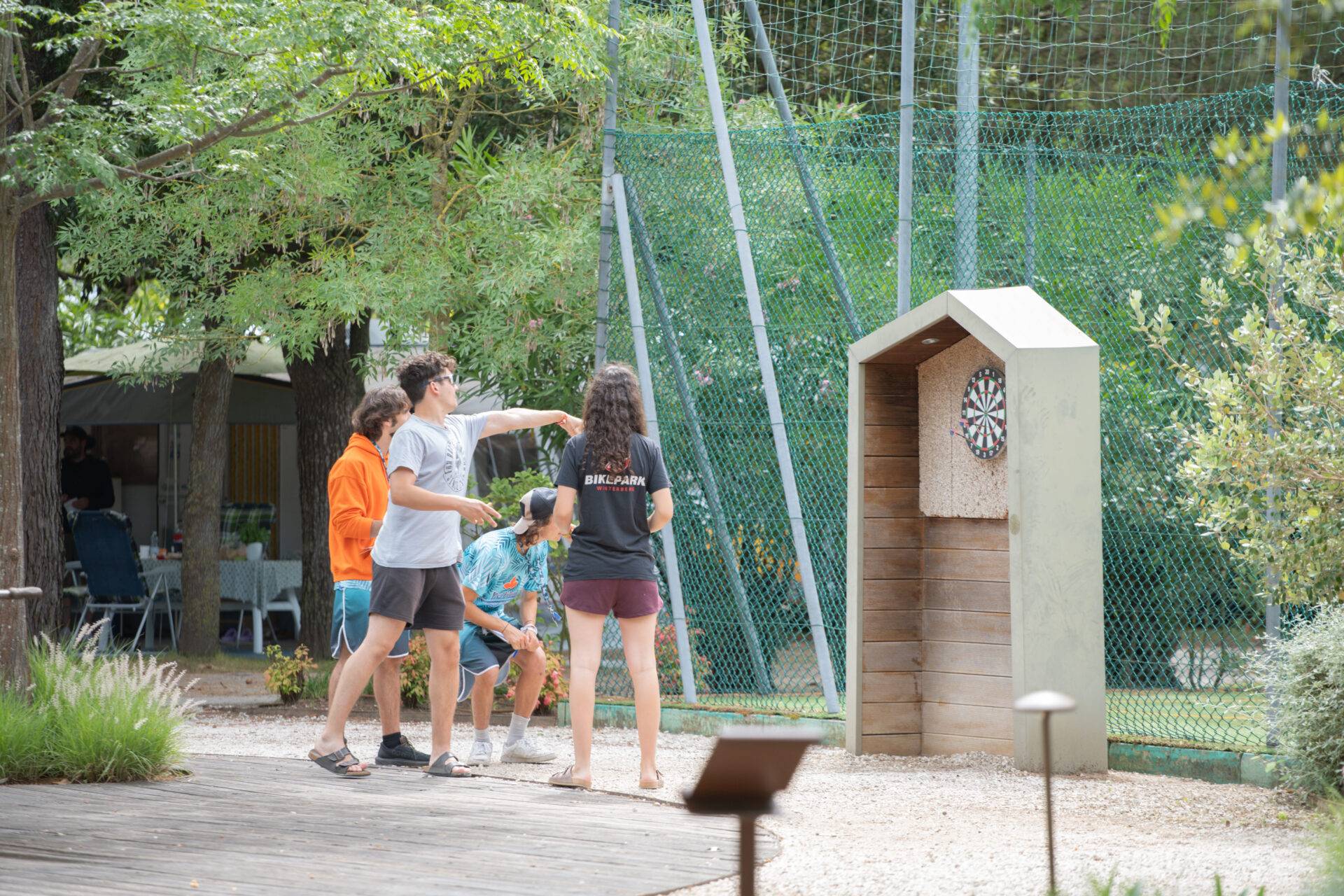 Gruppo di ragazzi gioca a freccette su un bersaglio all'aperto, tra alberi e vegetazione, durante l'animazione.