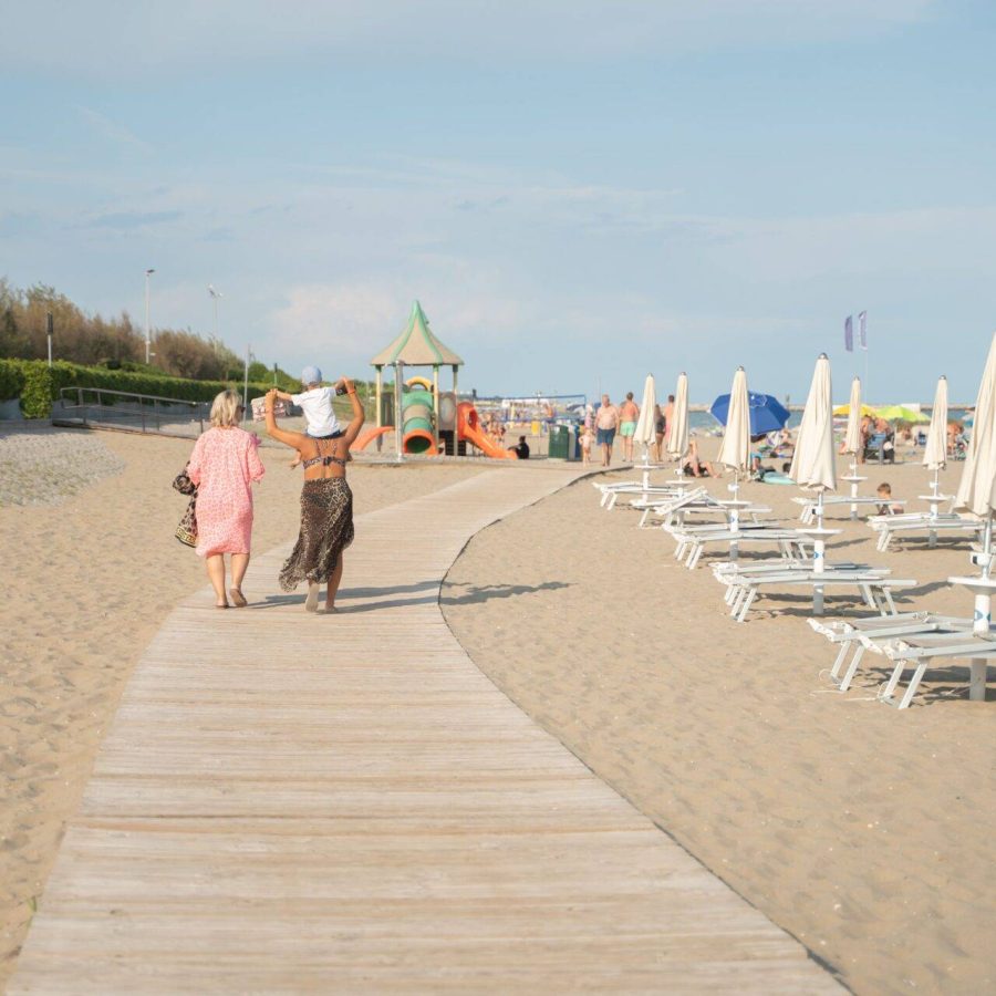 Passerella sulla sabbia verso la spiaggia attrezzata del Camping Dei Fiori, con ombrelloni, lettini e famiglie.